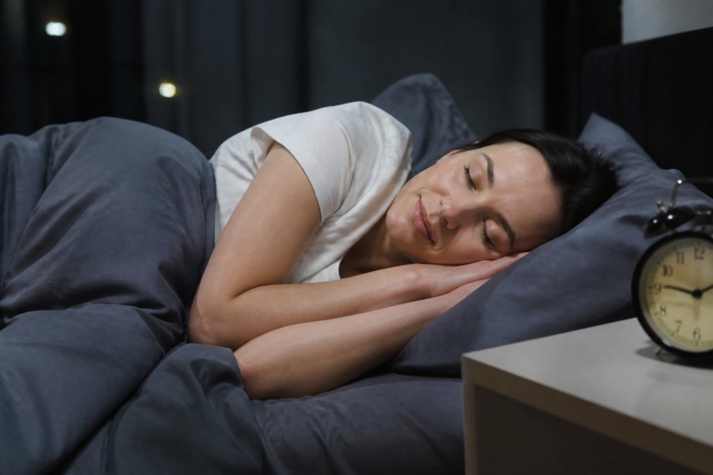 Person in a peaceful, darkened bedroom with natural soft lighting, lying in bed with a relaxed posture, quality bedding, and a calm environment