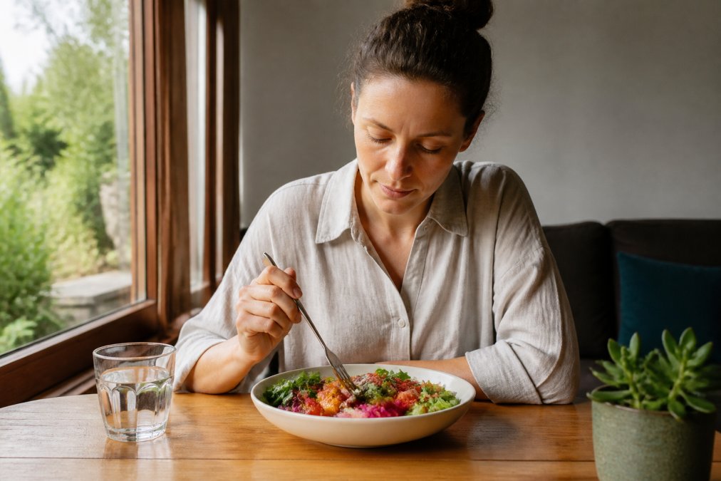 A person sitting at a wooden table, eating a colorful bowl of whole foods slowly with a fork, eyes focused on the meal, natural light from a window