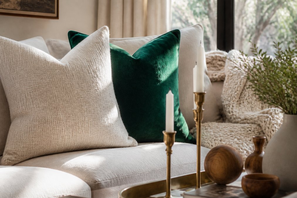 Close-up detail of a coordinated living room vignette showing a cream-colored linen throw pillow with subtle texture, a jewel-tone emerald velvet accent pillow, warm brass candlesticks, natural wood decorative objects, and layered texture from a chunky knit throw blanket, arranged on a neutral sofa in soft afternoon light