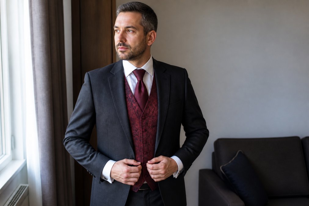 Professional man wearing a charcoal suit jacket with a burgundy paisley waistcoat, white dress shirt, coordinated burgundy silk tie, and silver cufflinks, photographed in natural daylight with soft window lighting