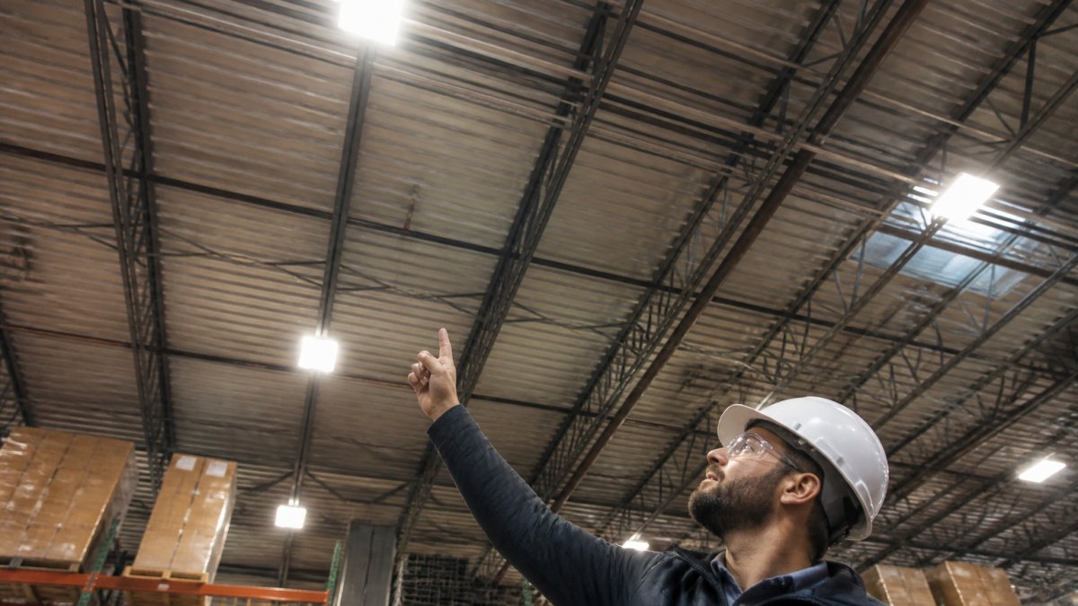 Professional facility manager or electrician inspecting high-bay warehouse lighting fixtures from below, pointing upward to assess existing conditions and fixture placement
