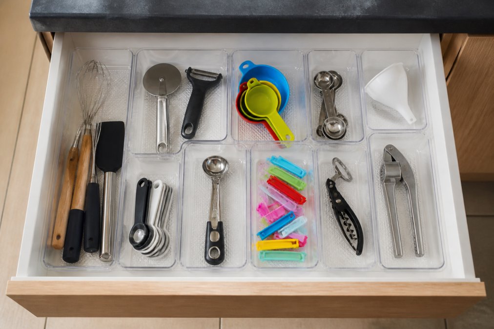 A neat kitchen drawer fitted with clear plastic organizers, holding utensils and small gadgets, shot from a top-down angle in natural light