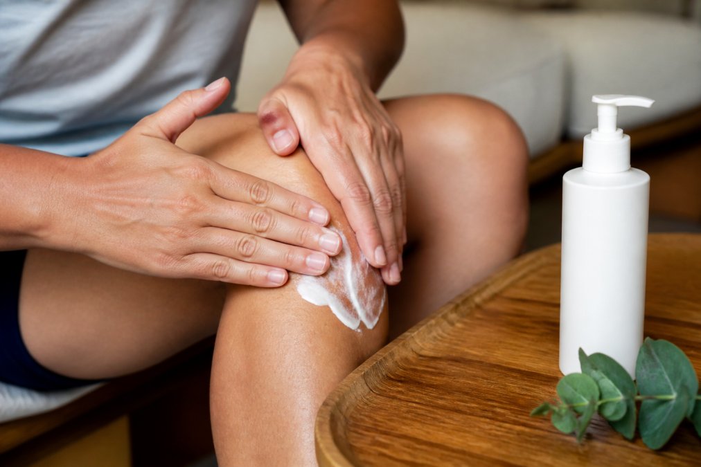 Close-up of a person's hands gently massaging a white lotion into their knee, with a white lotion bottle and a small green plant sprig on a wooden table nearby
