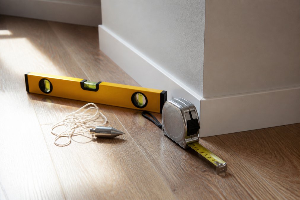 Close-up of a steel tape measure, spirit level, and plumb line arranged on a wooden floor beside an alcove wall, with natural daylight illuminating the measurement tools