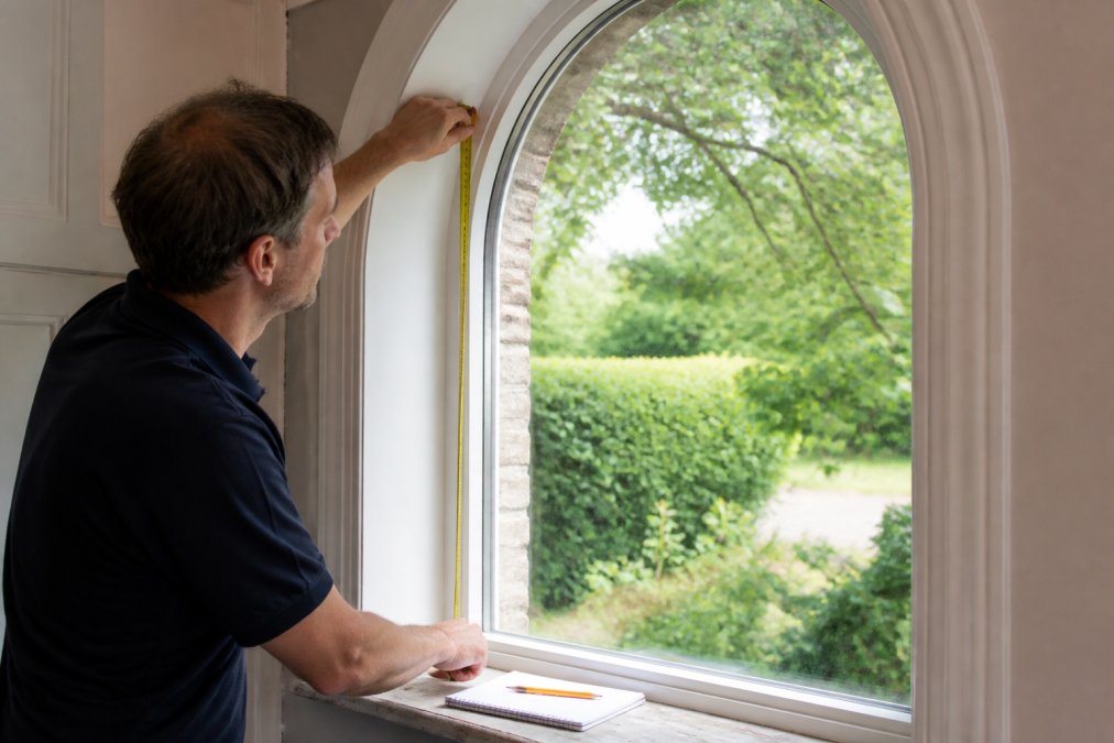 Close-up of a person holding a steel tape measure against the inside frame of a tall arched window in a period property, with natural light coming through the glass and a notepad and pencil resting on the windowsill