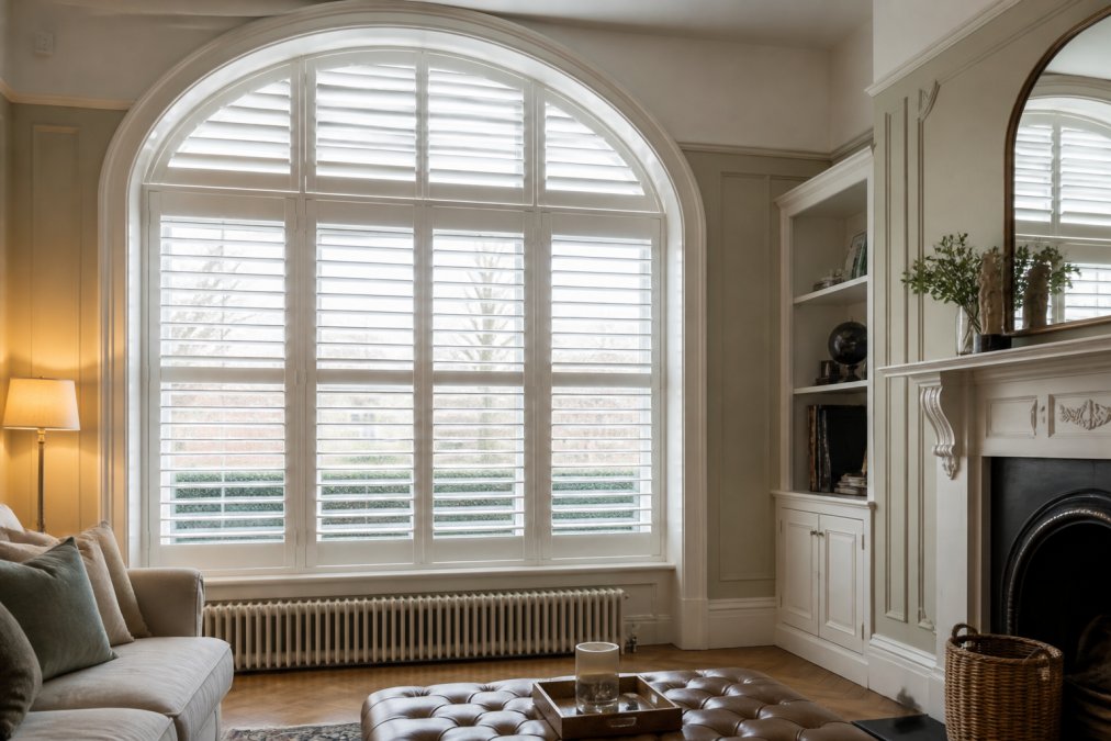 Elegant white plantation shutters fitted to a large arched window in a bright, classically decorated living room, with soft natural light filtering evenly through the louvres and a period fireplace visible in the background