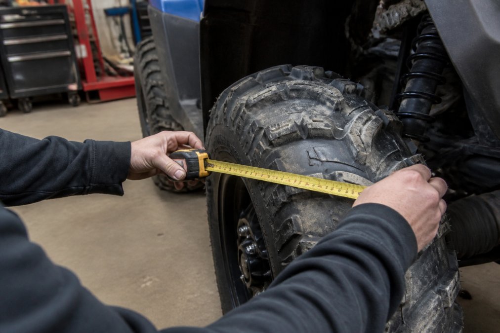 Hands holding a tape measure across a mounted tire to determine the correct ATV tire size in a garage.
