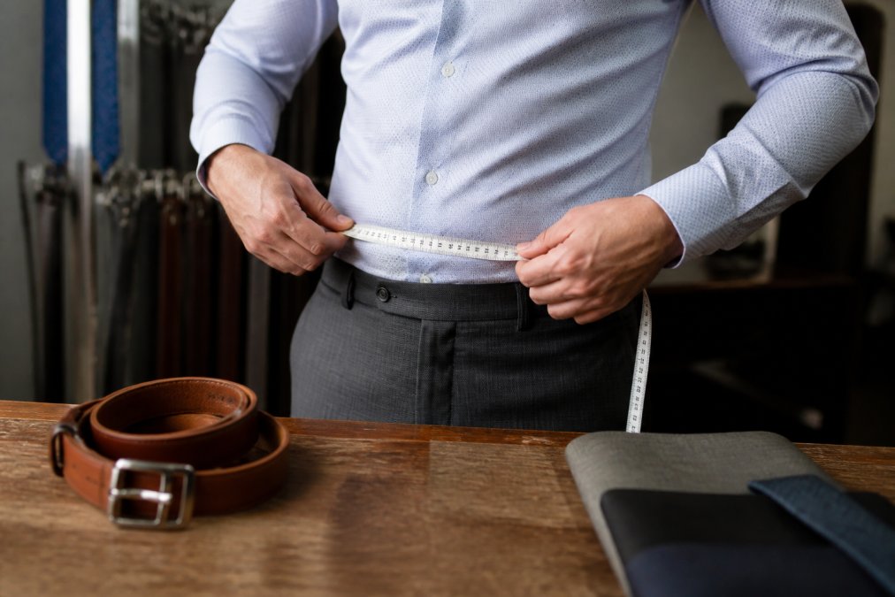 A close-up of a man's hands holding a flexible tape measure around his waist over a dress shirt, with a leather belt nearby on a wooden table