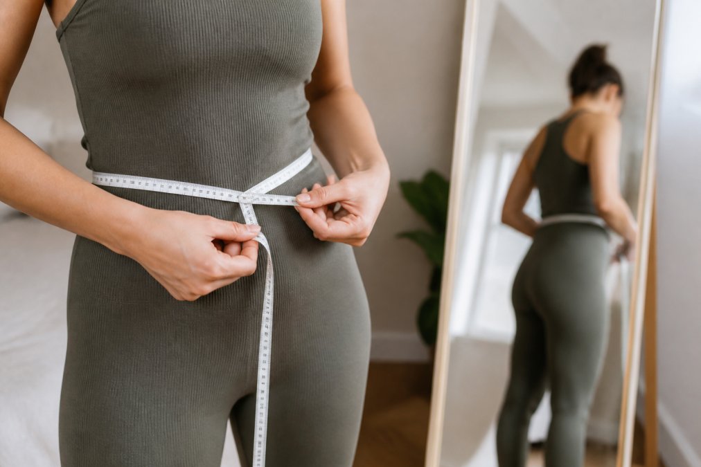Close-up of hands holding a flexible measuring tape against a body, showing proper tape tension and positioning over form-fitting clothing with a full-length mirror visible in the background