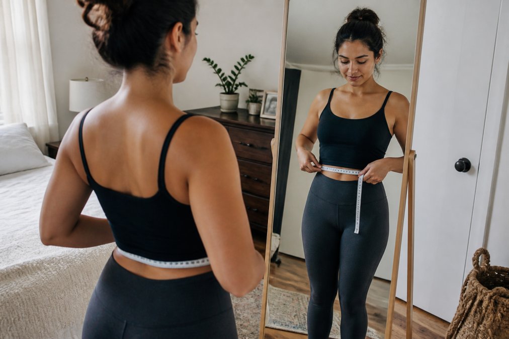 A woman standing in front of a full-length mirror in a bright bedroom, holding a soft measuring tape around her natural waist, wearing fitted everyday clothes