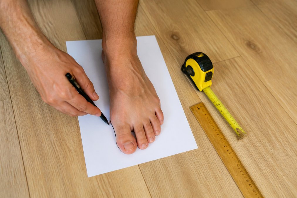 A person's bare foot being traced on white paper with a pen, a tape measure and wooden ruler resting nearby on a light hardwood floor