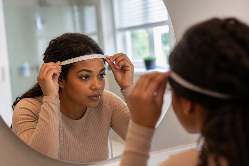 Close-up of a woman's hands holding a flexible tape measure around her head just above the ears, looking into a bathroom mirror with soft natural light