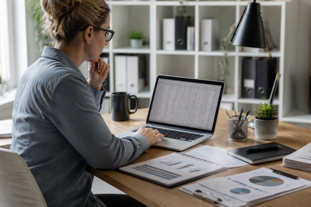 Professional marketing manager reviewing influencer analytics dashboard on laptop screen in modern office, with campaign spreadsheets and creator data visible on desk