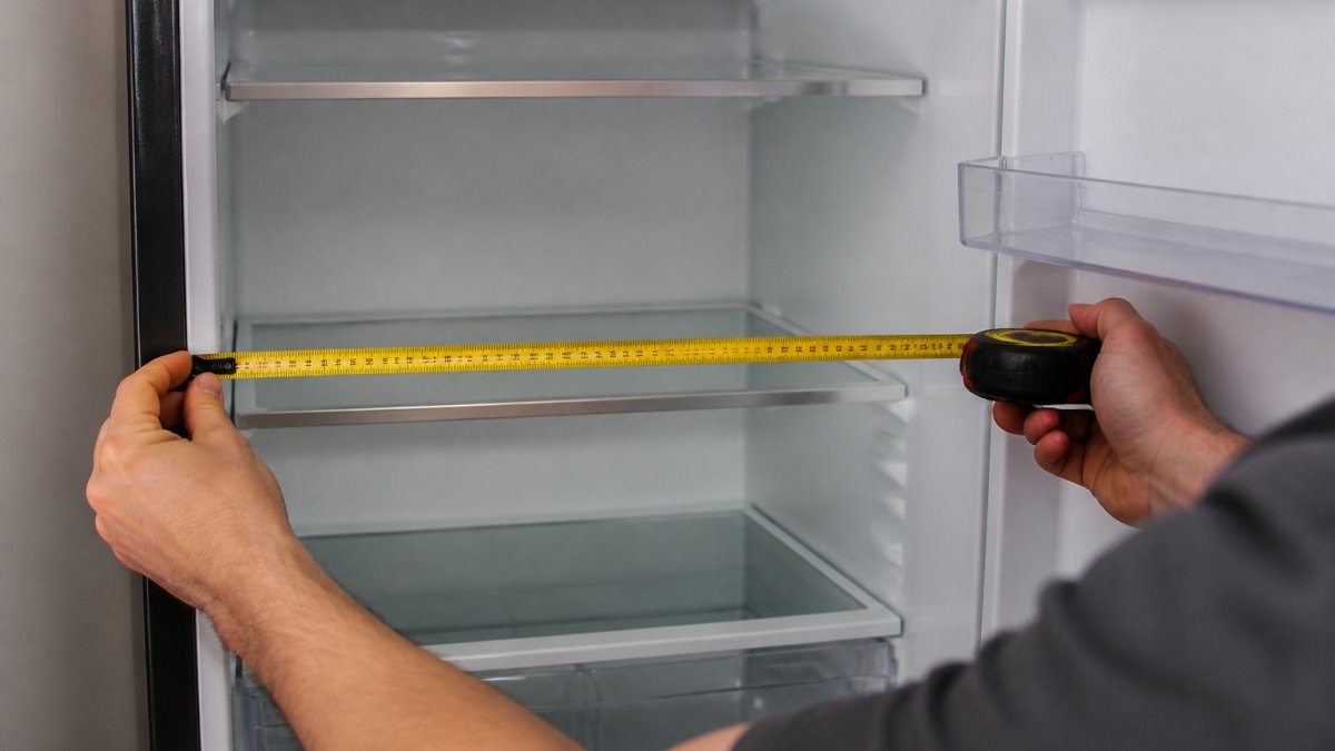 Close-up of hands using a metal tape measure inside an open refrigerator, measuring the interior width with focus on the measurement markings and refrigerator shelving visible in background
