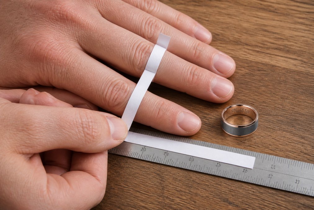 Close-up of a person's hand with a narrow strip of white paper wrapped around the base of the ring finger, with a ruler placed alongside to measure the length in millimetres on a wooden surface