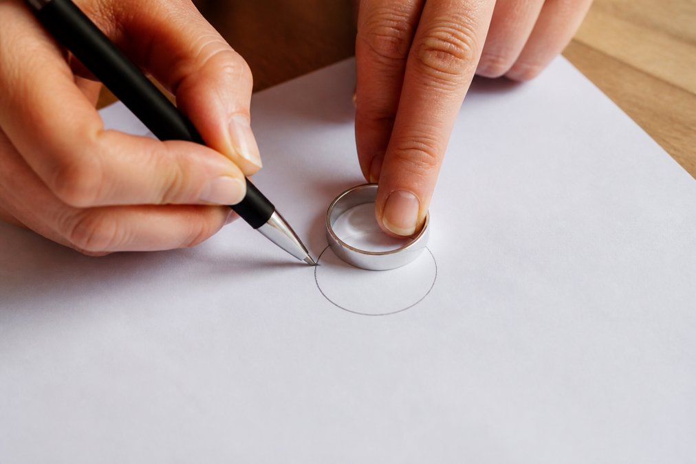 Close-up of a hand carefully tracing a silver ring onto white paper with a pen, showing the circular outline clearly visible