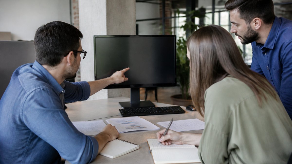 Professional team reviewing performance metrics and baseline data on desktop monitor in modern office, with notebooks and data sheets visible on desk