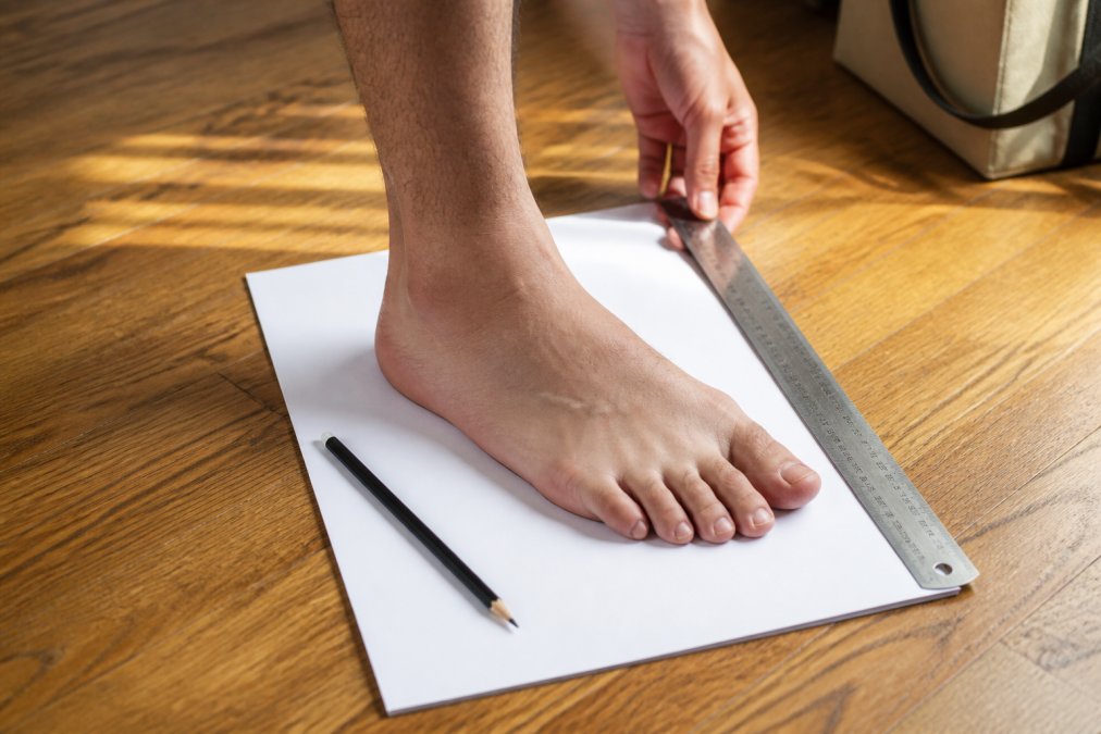 Close-up of a person's bare foot placed flat on a white sheet of paper on a hardwood floor, with a ruler alongside the foot tracing and a pencil nearby, shot in bright natural daylight
