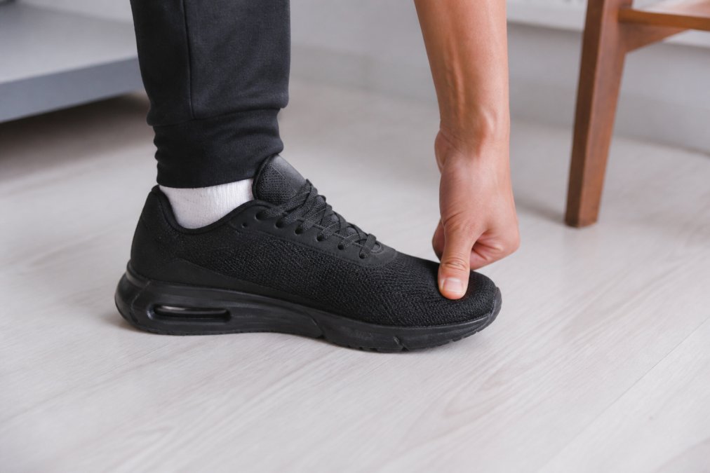 Person standing on a clean light-colored floor, pressing their thumb against the toe box of a black sneaker to check for proper toe room, shot from a low angle with soft studio lighting