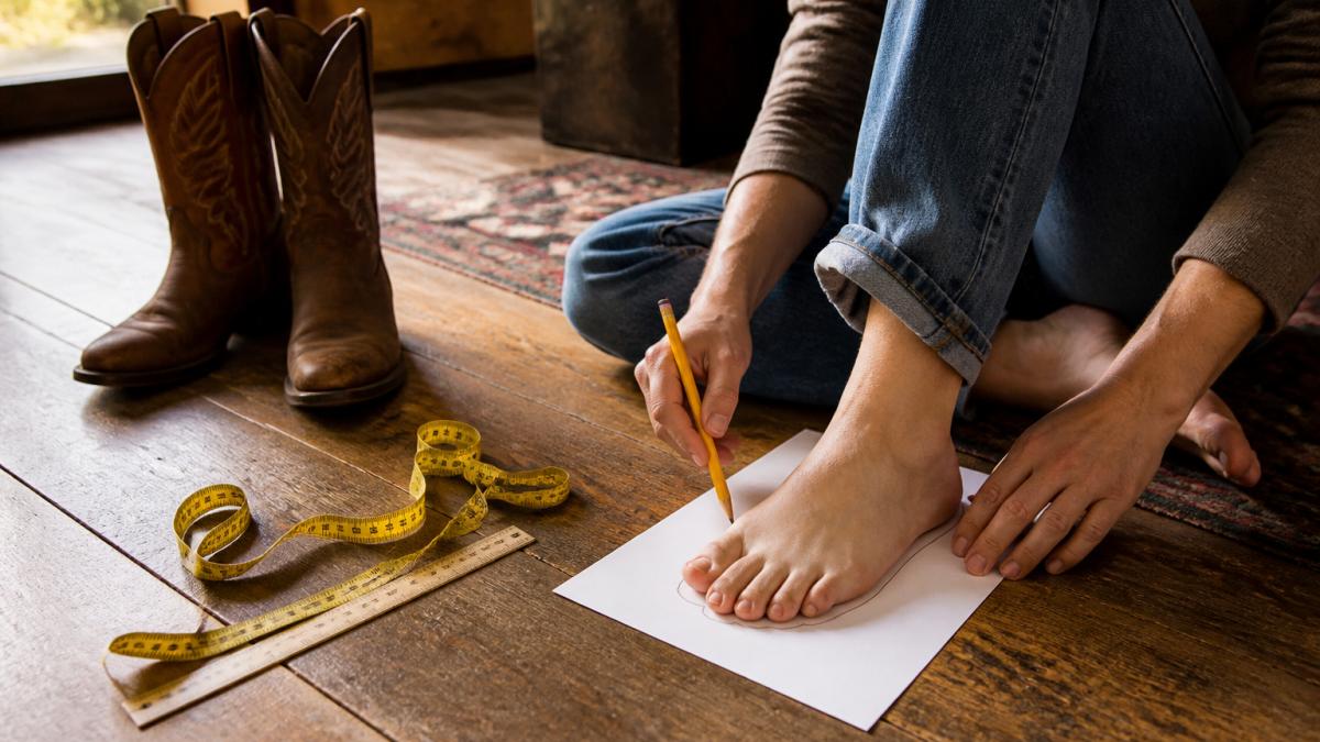 A person sitting on a wooden floor tracing their bare foot on a sheet of paper with a pencil, a fabric measuring tape and a ruler nearby, warm natural light from a window, casual western home setting