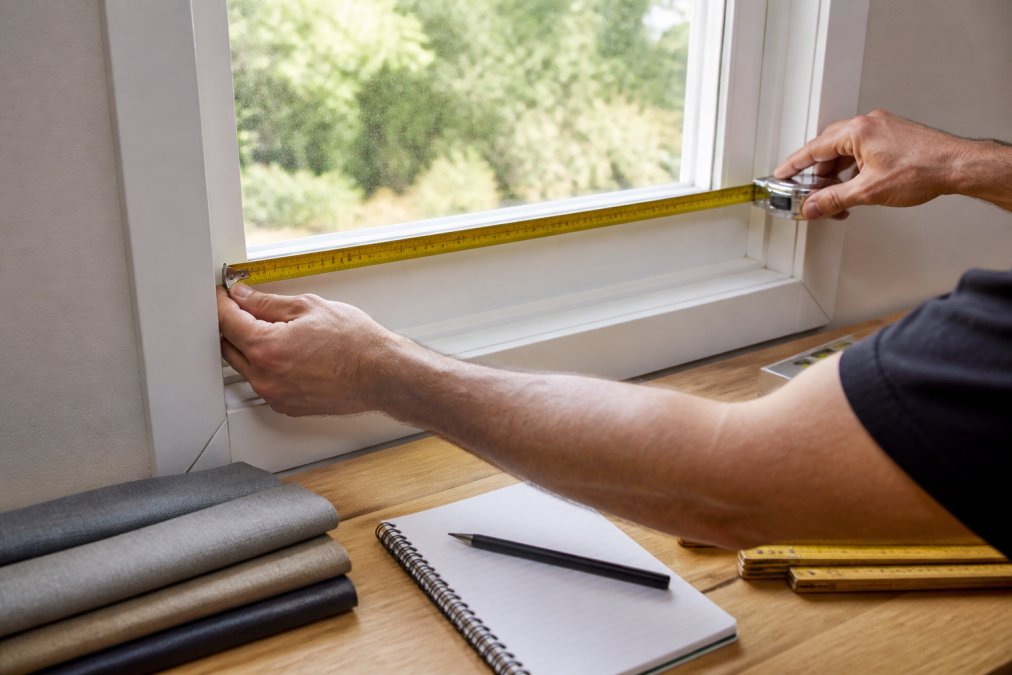 Close-up of a person's hands holding a metal tape measure horizontally against a window frame, measuring the width of the glass opening with natural daylight streaming through the window