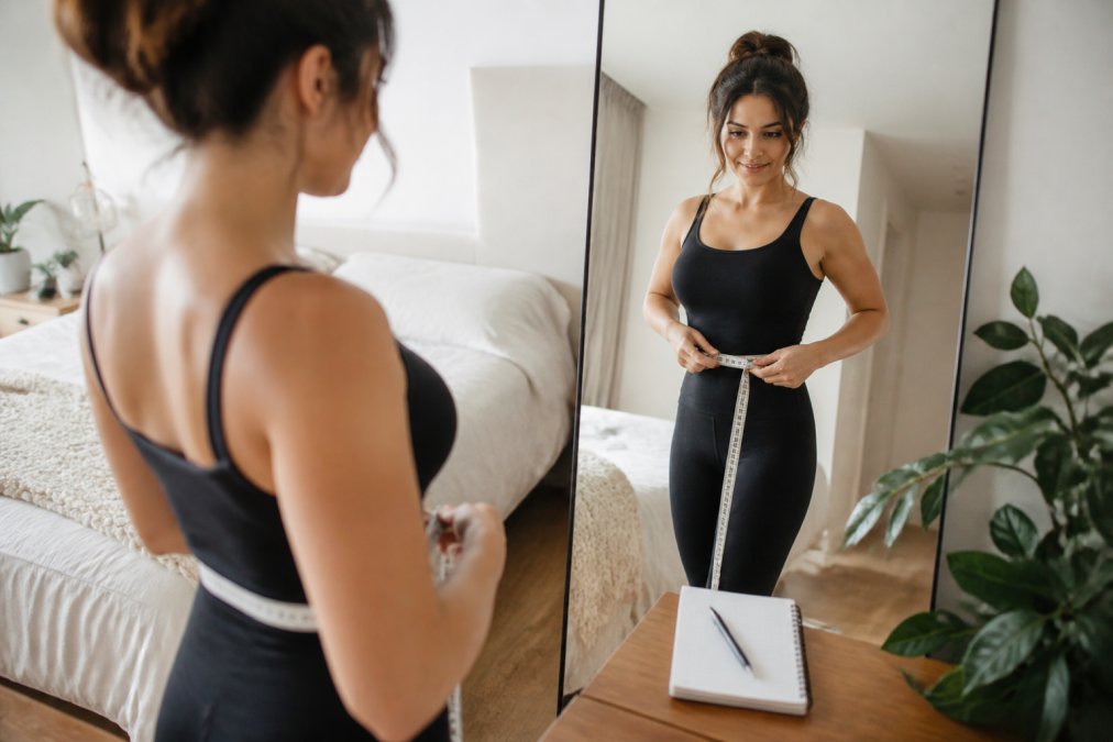 A woman standing in front of a full-length mirror in a bright bedroom, holding a soft measuring tape around her waist, wearing fitted clothing, with a notepad nearby for recording measurements