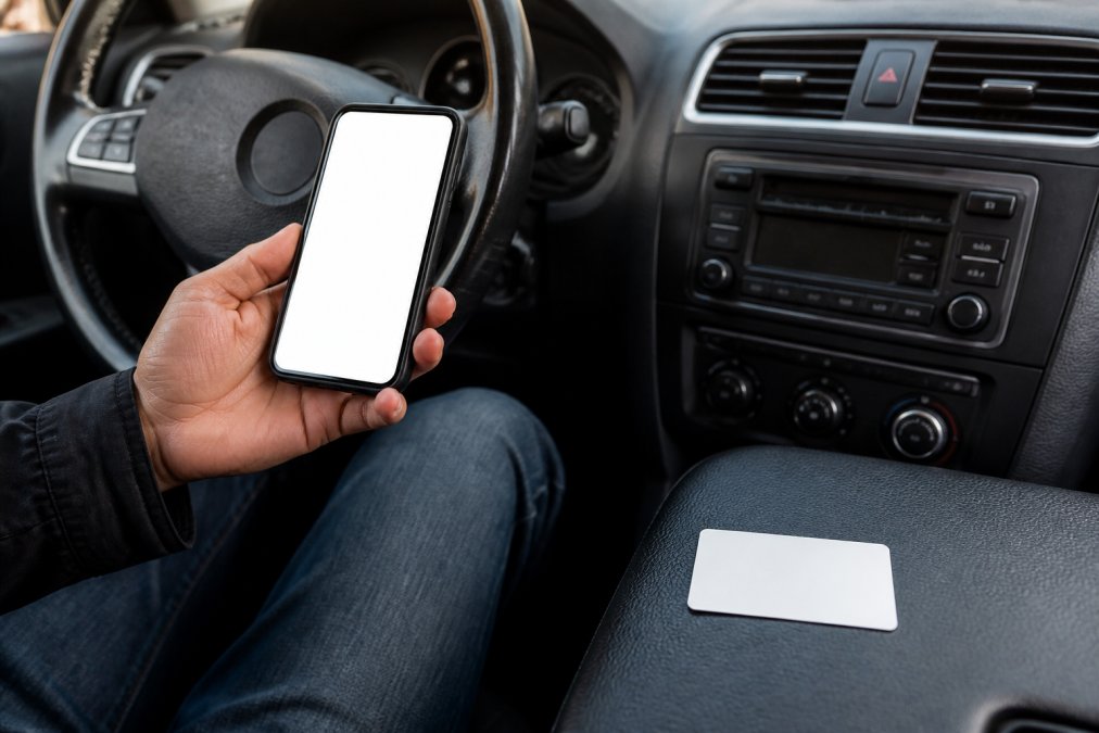 A driver holding a smartphone displaying a digital insurance card while sitting in a parked car, with a physical insurance card visible on the dashboard