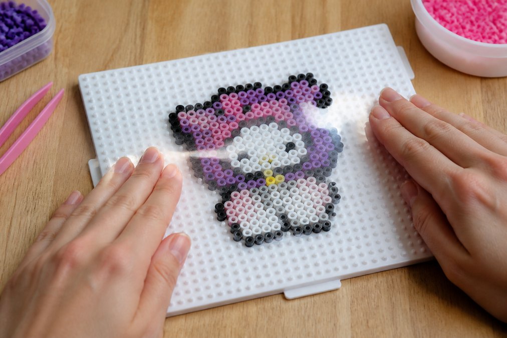 A pair of hands carefully pressing a strip of clear tape across a finished fuse bead pixel art design on a white pegboard, smoothing it flat to hold the beads in position before lifting, photographed on a clean wooden table with soft natural light