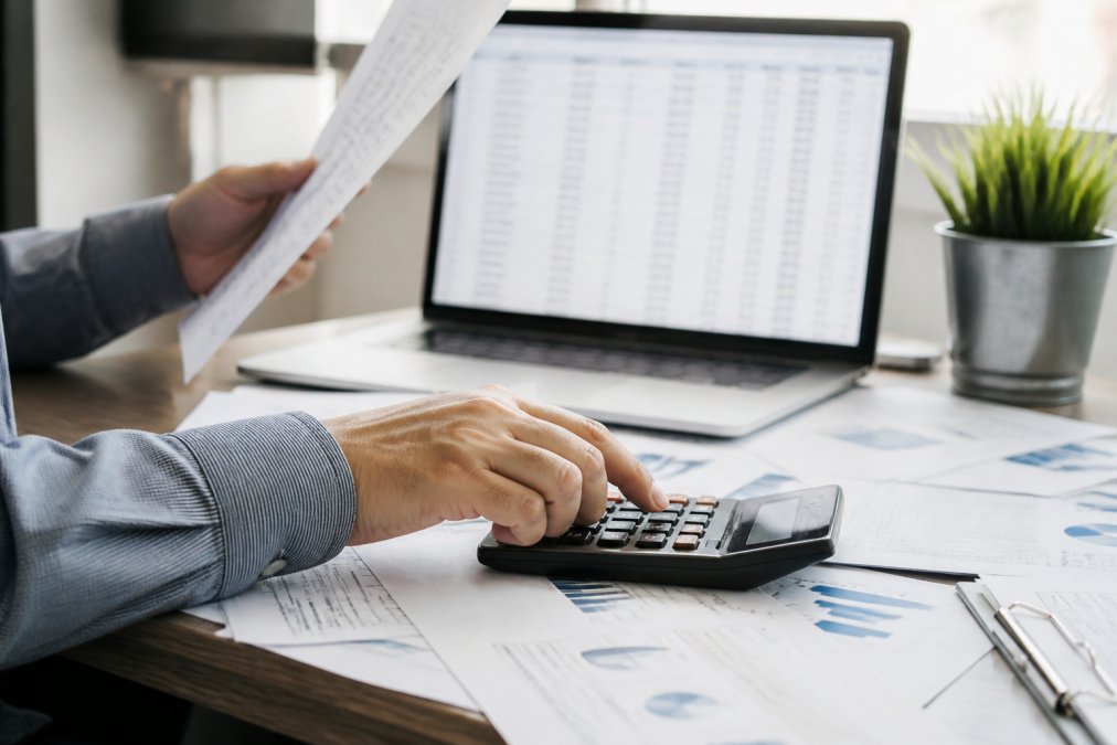 Close-up of a real estate investor's hands reviewing financial documents with a calculator at a desk, laptop showing a spreadsheet in the background, natural office lighting