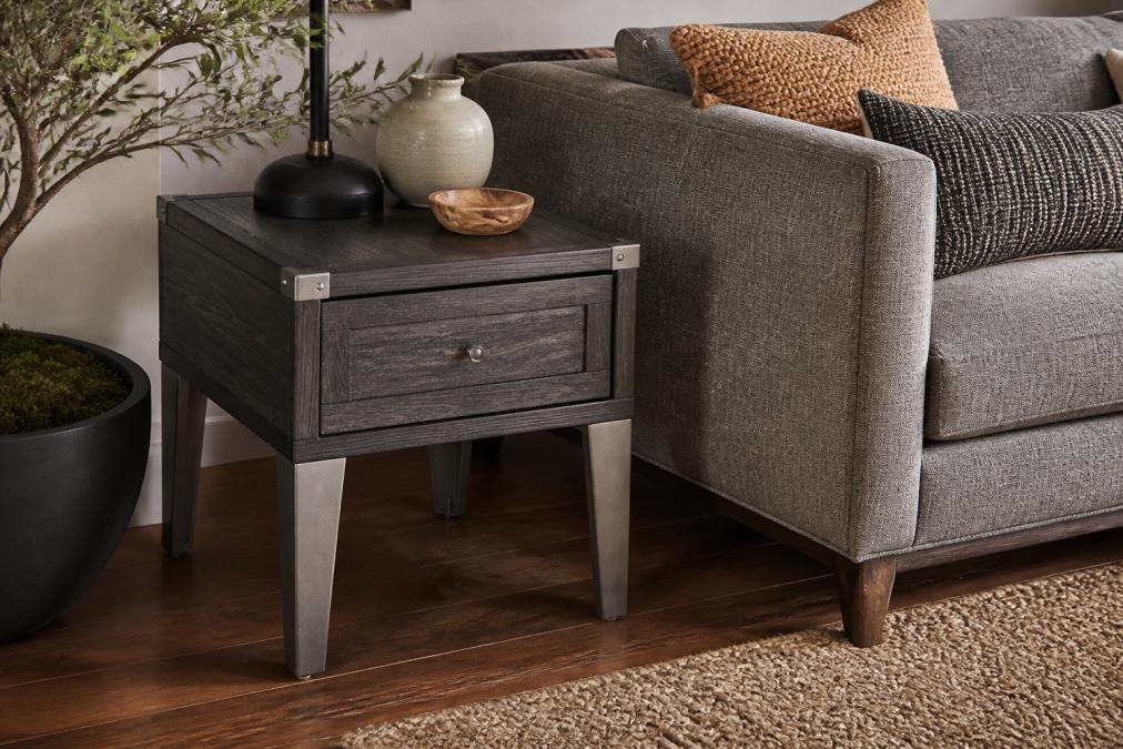 A well-styled living room corner featuring a rustic-industrial end table with mixed wood tones and metal accents positioned next to a contemporary gray upholstered sofa, with warm wooden flooring and layered textures creating a harmonious blend of styles