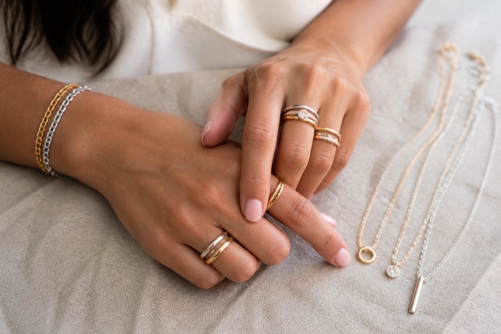 Woman wearing stacked rings and layered necklaces to mix gold and silver jewelry on a neutral fabric background.