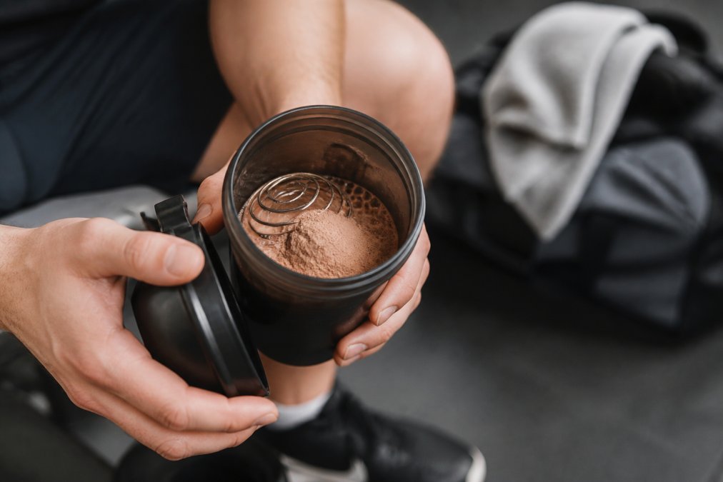 Close-up of hands holding a shaker cup with a wire whisk ball, a scoop of chocolate plant protein powder sitting on top of liquid, with a gym bag and towel blurred in the background