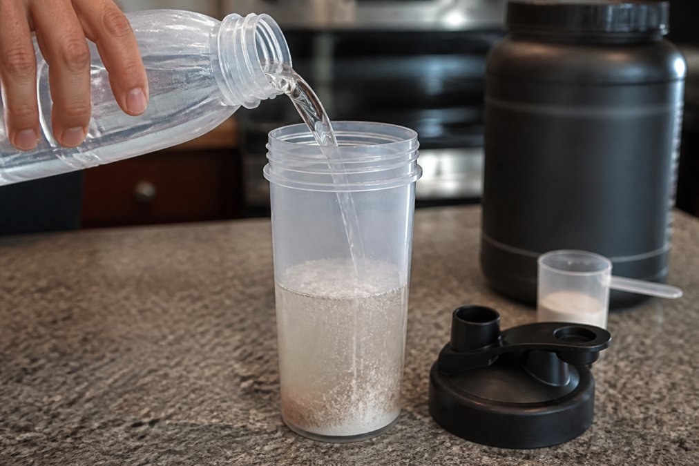 Close-up of a hand pouring water into a clear shaker bottle that already contains pre-workout powder, showing the powder fully submerged in the liquid before shaking