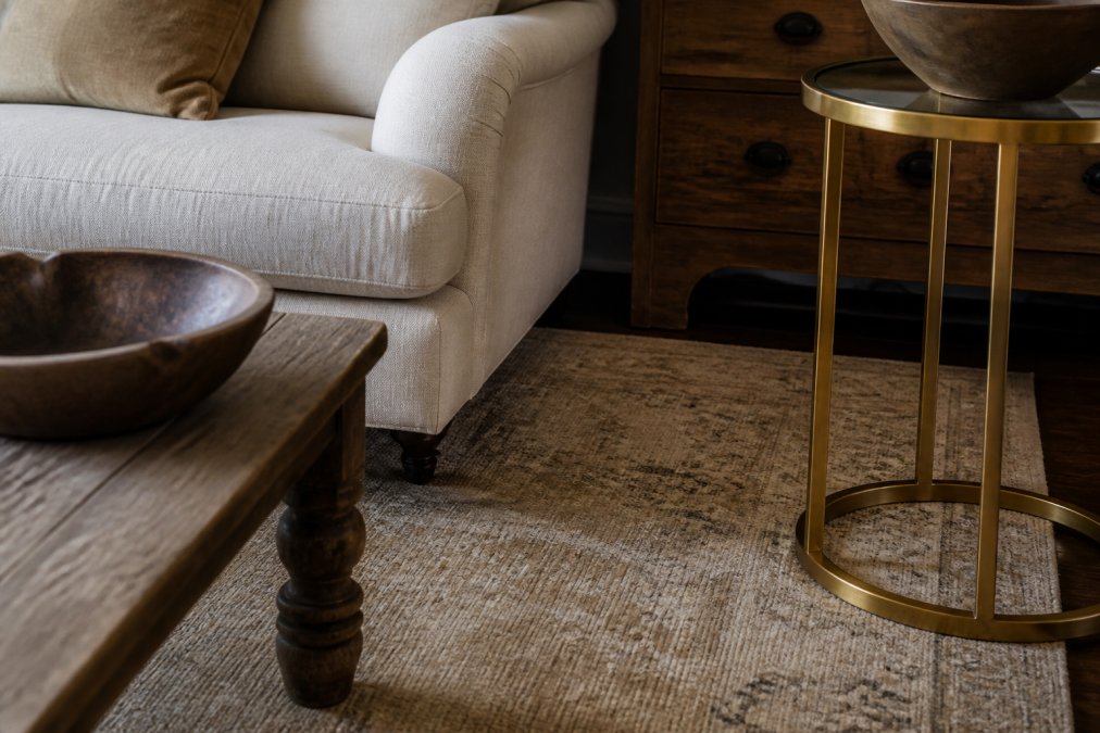 Close-up detail of a living room corner showing layered textures including a distressed vintage beige area rug with subtle pattern, soft linen upholstery in warm cream, vintage wood furniture with visible grain and patina, polished gold metal accents on a contemporary side table, and natural wood tones creating rich textural contrast