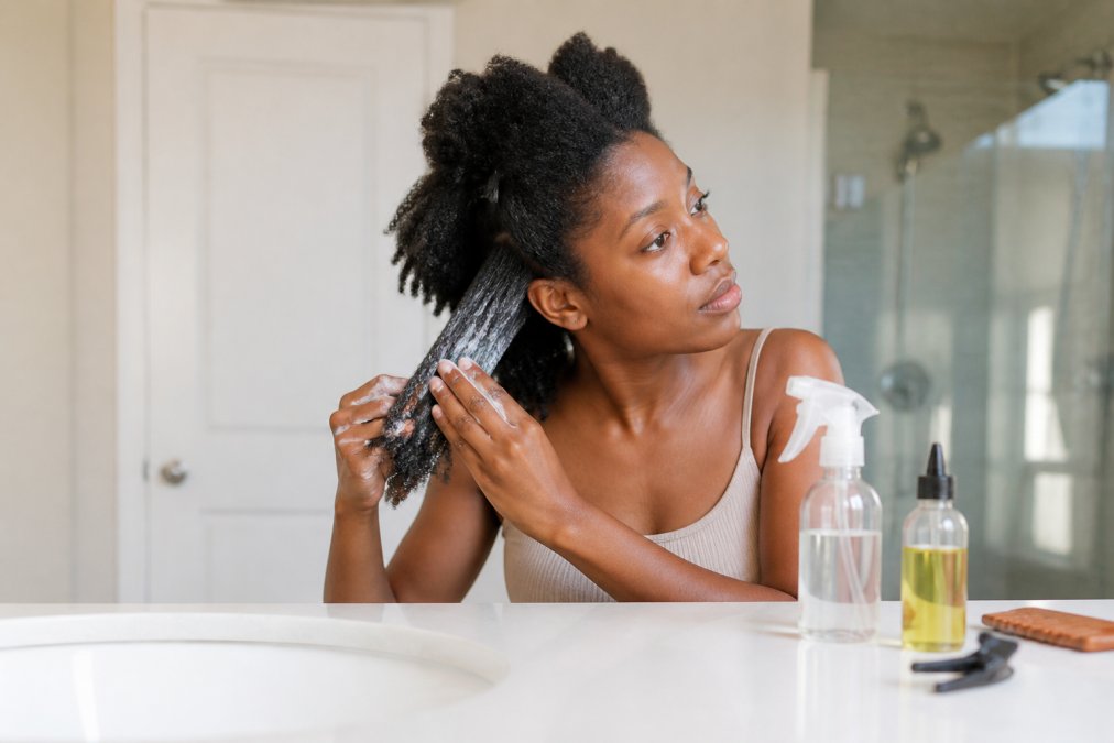 A woman with 4C coils in a bright bathroom applying leave-in conditioner to sections of her hair, with a spray bottle of water and a small bottle of oil on the counter beside her