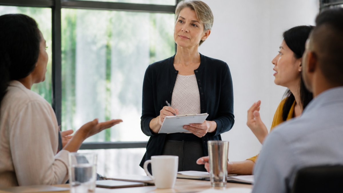 A woman leader in her fifties standing calmly before a small team in a bright meeting room, listening attentively with a notepad in hand while colleagues speak