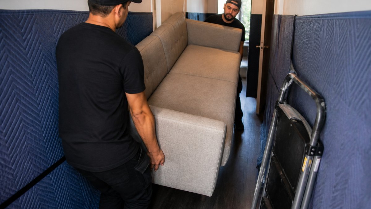 Two movers carefully navigating a large sofa through a narrow apartment hallway with protective padding on the walls and a moving dolly nearby