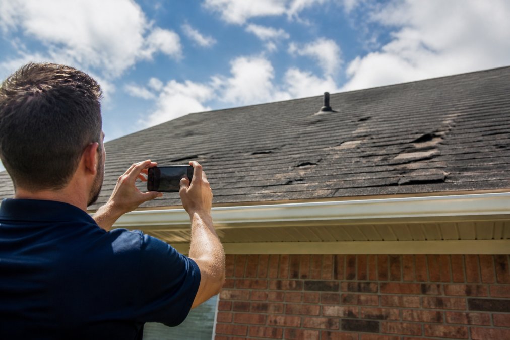 Homeowner photographing roof damage with smartphone from ground level, capturing visible hail damage and missing shingles with clear detail and natural daylight
