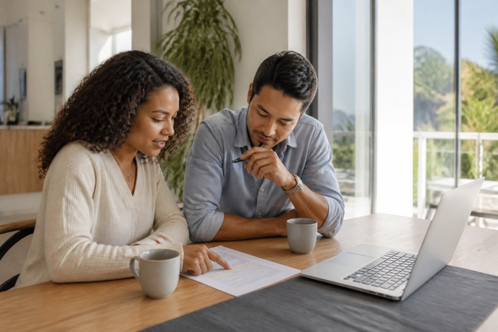 Two people reviewing a printed lease agreement and a laptop at a bright dining table, one pointing at a highlighted clause, coffee mugs nearby, relaxed professional setting