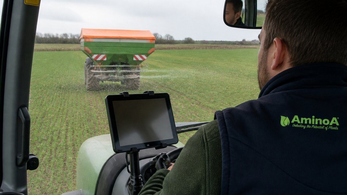 A tractor with a spinner spreader applying fertiliser across a field, with a tablet controller mounted in the cab showing a field map, photographed in flat overcast daylight