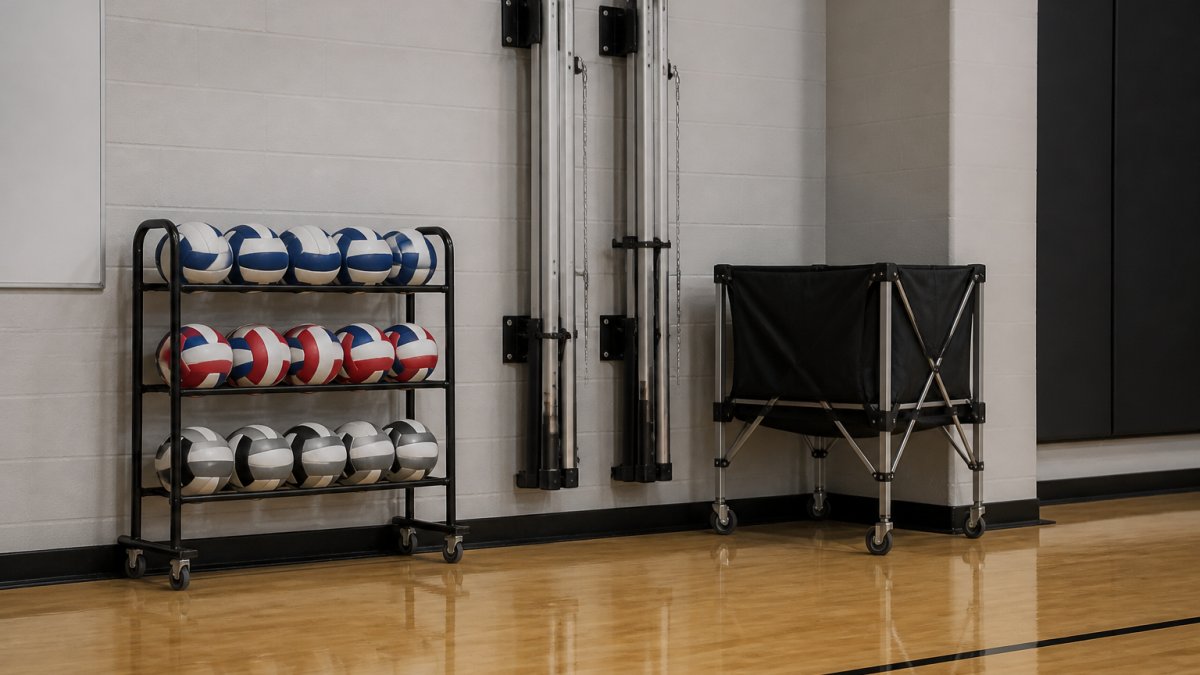 A gym storage room with a three-tier volleyball ball rack holding a dozen balls, wall-mounted vertical brackets holding collapsed net posts, and a rolling transporter cart parked against the wall, with clear floor space visible