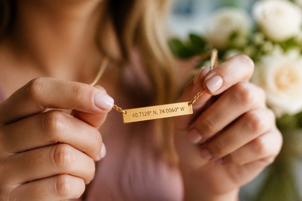 Close-up of a woman's hands holding a delicate gold bar necklace with engraved coordinates, soft natural light, blurred romantic background