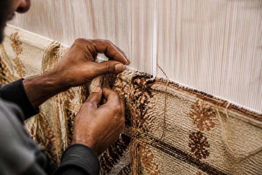 Close-up of artisan hands working on a hand-knotted rug loom, fingers positioning individual knots with natural wool fibers, traditional weaving technique visible with warm studio lighting