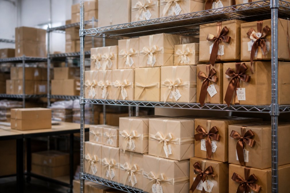 Neatly stacked boxes of wrapped personalized gifts arranged on metal shelving in a well-organized fulfillment area, with custom labels clearly visible on each package under bright warehouse lighting