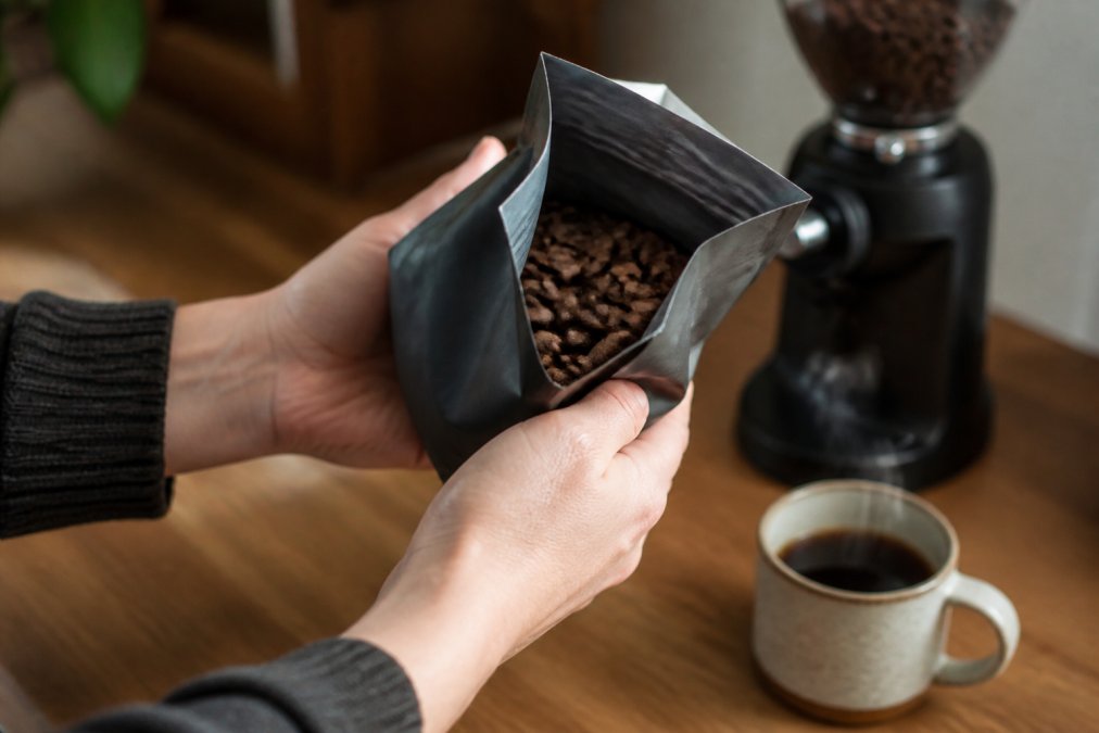 Close-up of a person's hands holding an open bag of whole bean coffee, tilting it to read the printed roast date label, with a steaming ceramic coffee cup and a burr grinder blurred in the background on a wooden kitchen counter