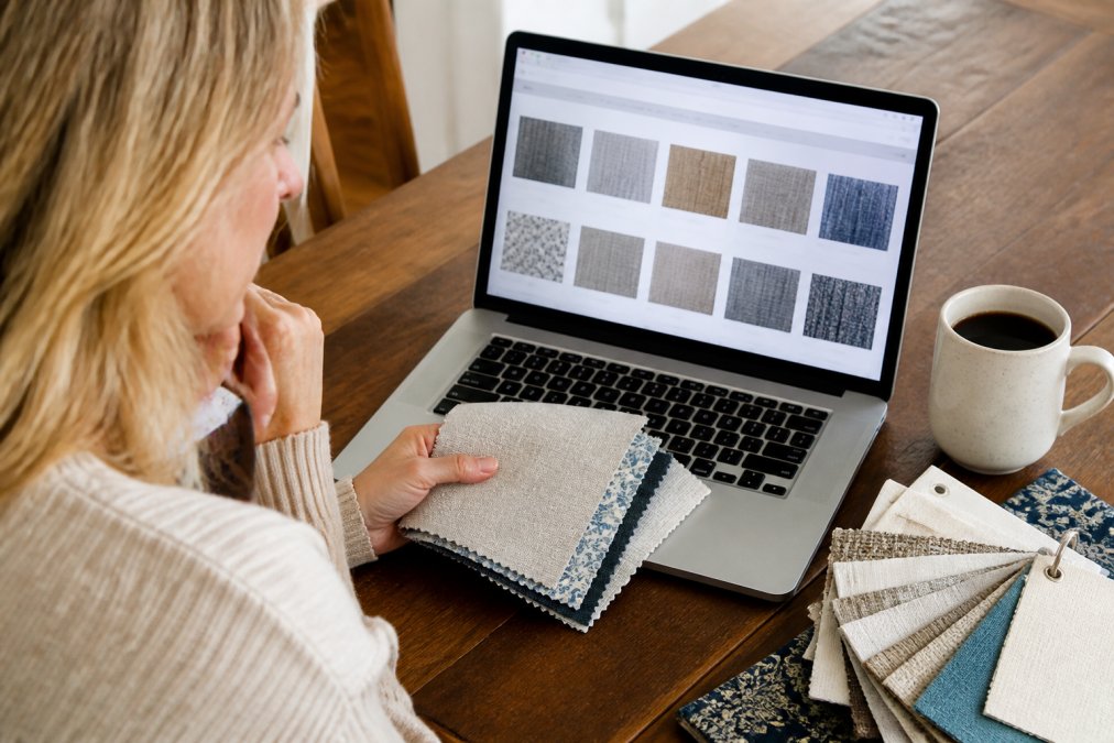 Fabric swatches fanned out next to a coffee mug on a wooden desk with an open laptop screen