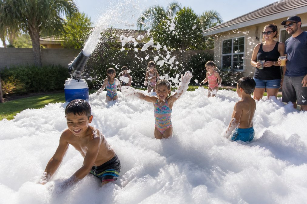 Children and adults playing in white foam at a backyard foam party, with a foam machine visible in the background and adults supervising from the side, natural afternoon sunlight
