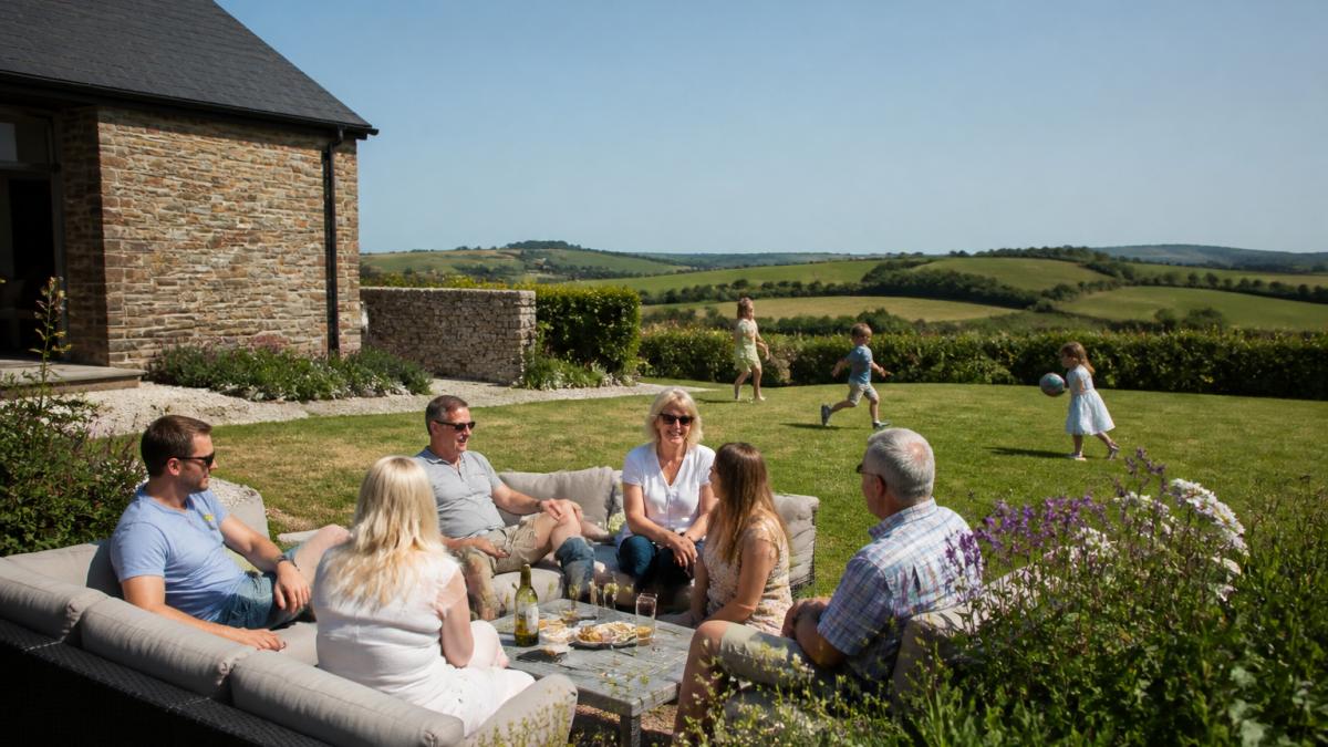 A multi-generational family group relaxing together in the enclosed garden of a converted stone barn on a sunny afternoon, with countryside views beyond and children playing on the lawn