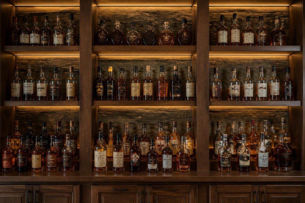 A well-organized home whiskey cabinet with bottles displayed upright on wooden shelves with warm accent lighting, mix of sealed and opened bottles arranged by region and age, with visible labels and organized spacing