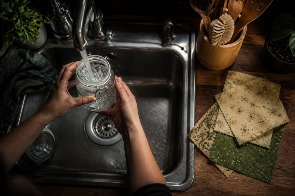 Overhead shot of hands rinsing and refilling a repurposed glass jar at a small kitchen sink, with beeswax wraps and a bamboo utensil holder on the counter beside it